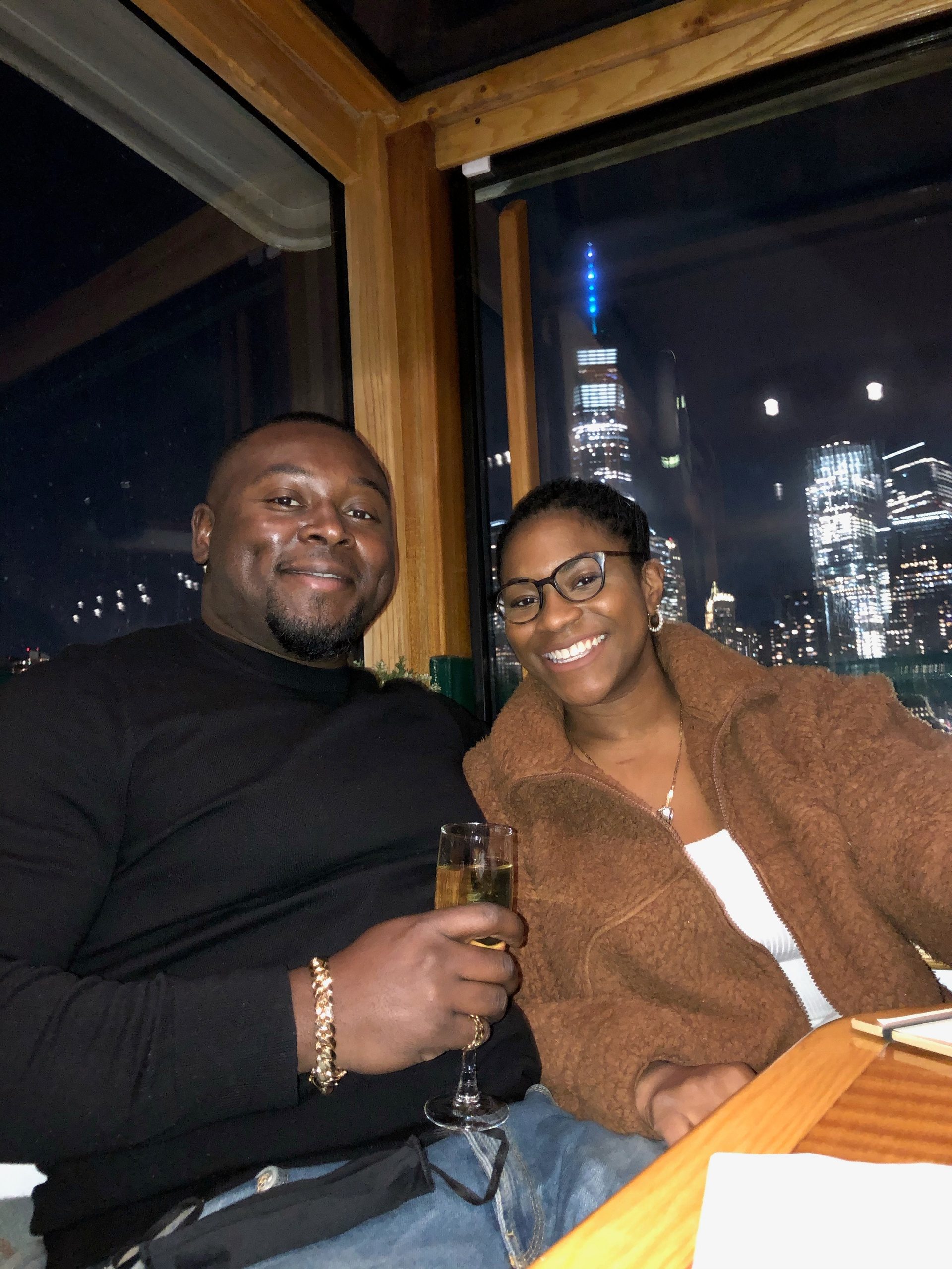 Two people smiling, seated in a dimly-lit restaurant with a city skyline visible through the window.
