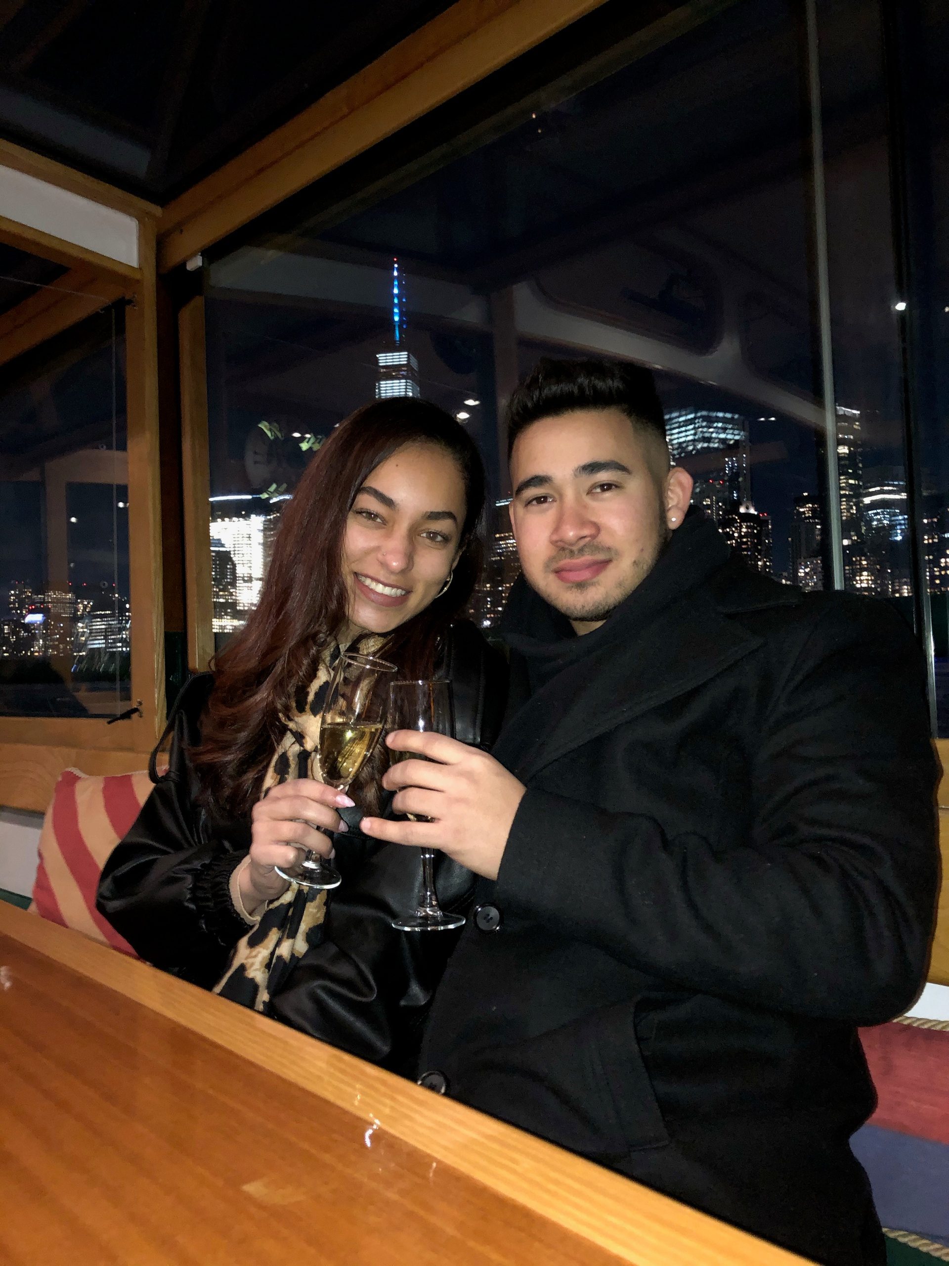 Two people seated indoors holding drinks, city lights visible through window.