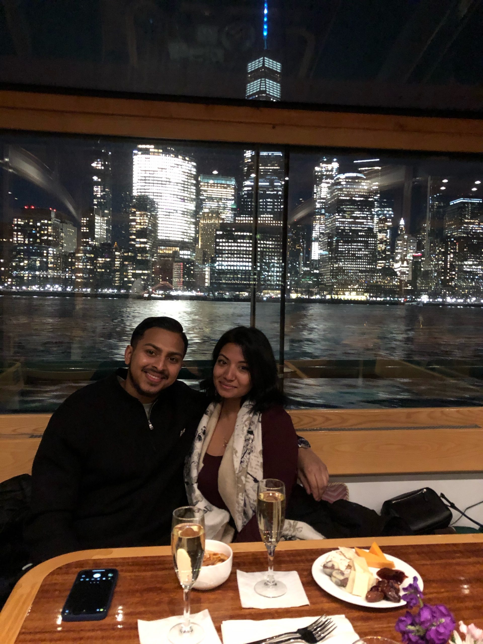 Couple dining with city skyline view, champagne, and dessert on a night cruise.