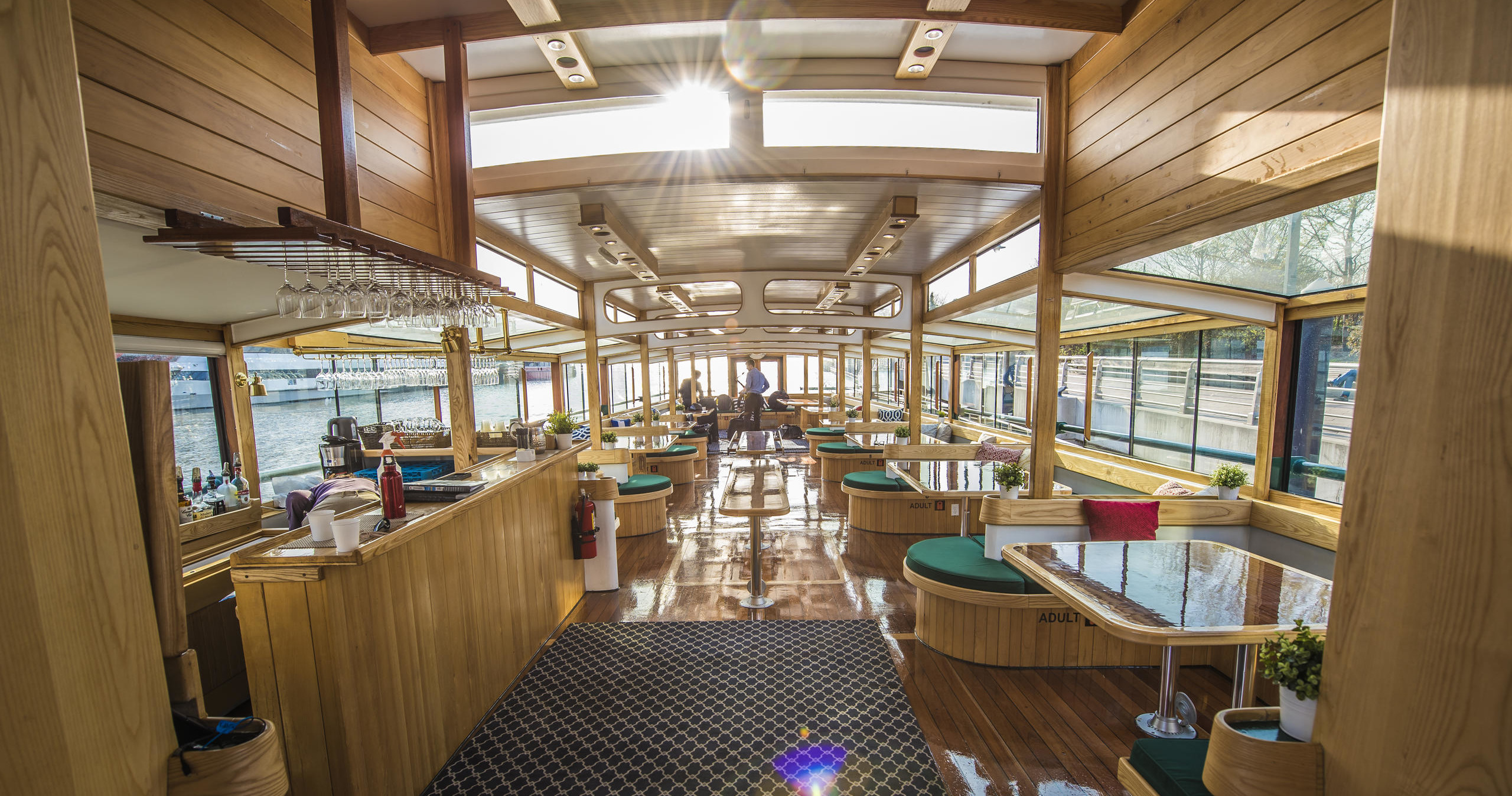 Spacious boat interior with wooden decor and tables, sunlight streaming through large windows.