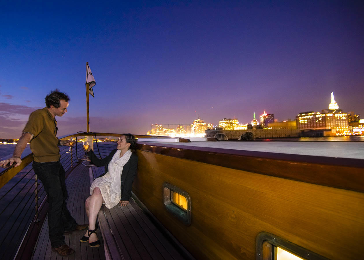 Two people chatting on a yacht deck with city lights in the background at dusk.