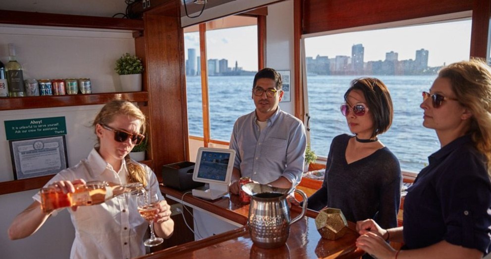 Bartender pouring drinks for three people on a boat with a city skyline view.
