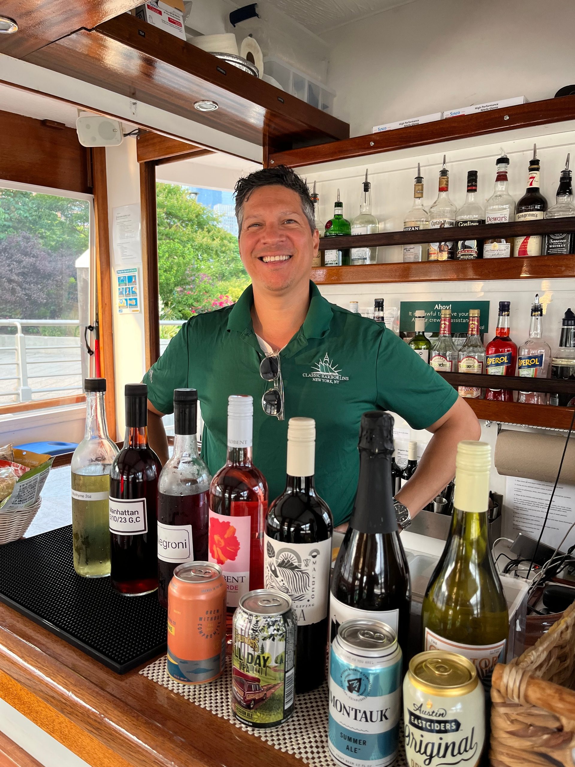 Smiling person behind a bar counter displaying various drinks and bottles.