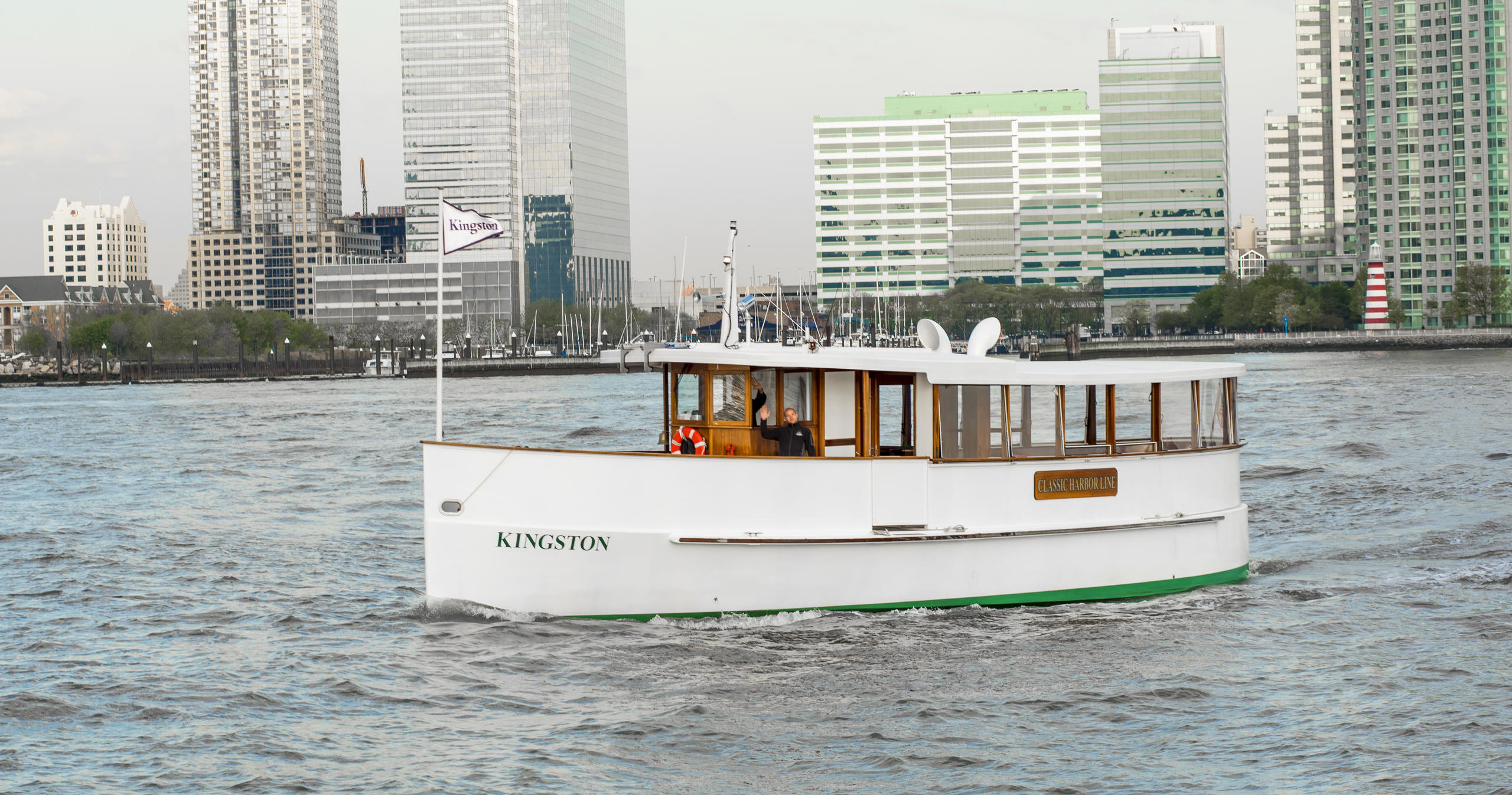 Small white boat named Kingston on a river with city buildings in the background.