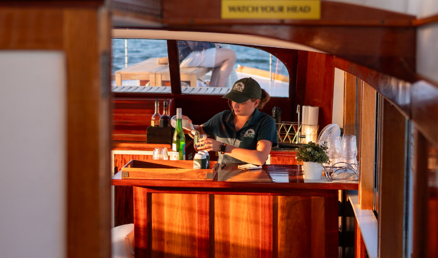 Person behind bar on a boat, pouring a drink with sunlight streaming through.