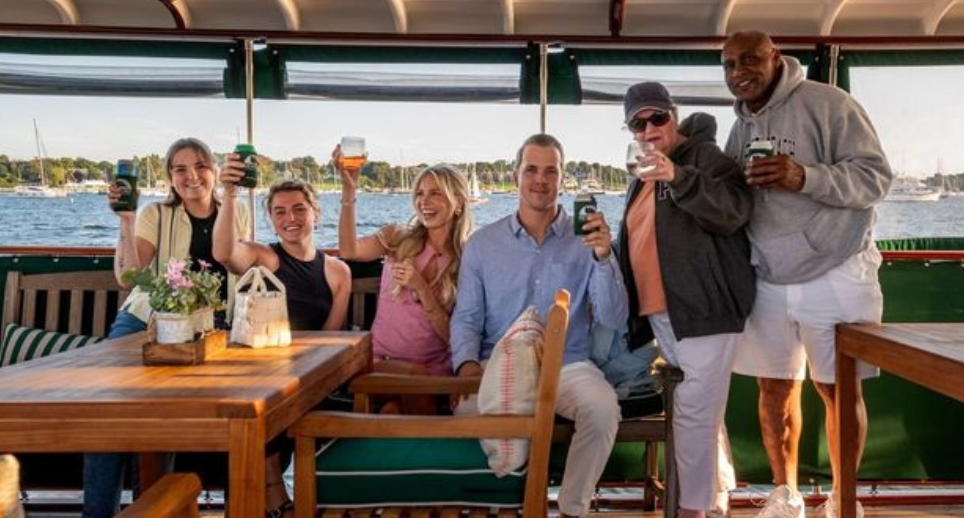 Group of six people smiling and holding drinks on a boat with water in the background.