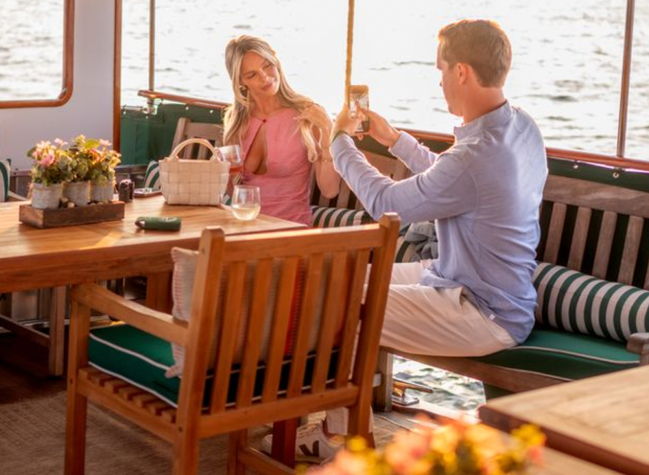 Couple toasting drinks on a boat with wooden furniture and water view.