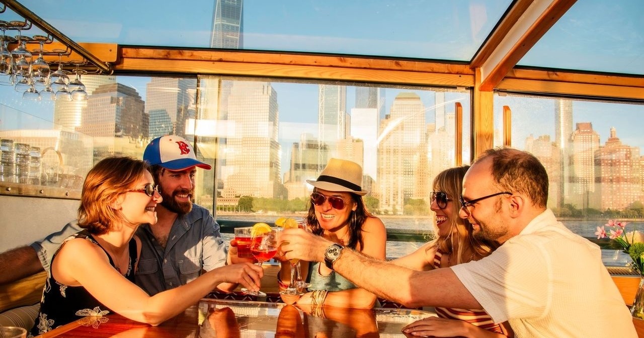 Group of five friends toasting drinks on a boat with city skyline view.