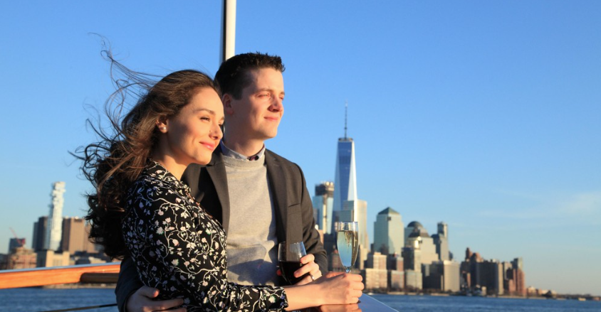 Couple on a boat, city skyline in background, holding drinks.
