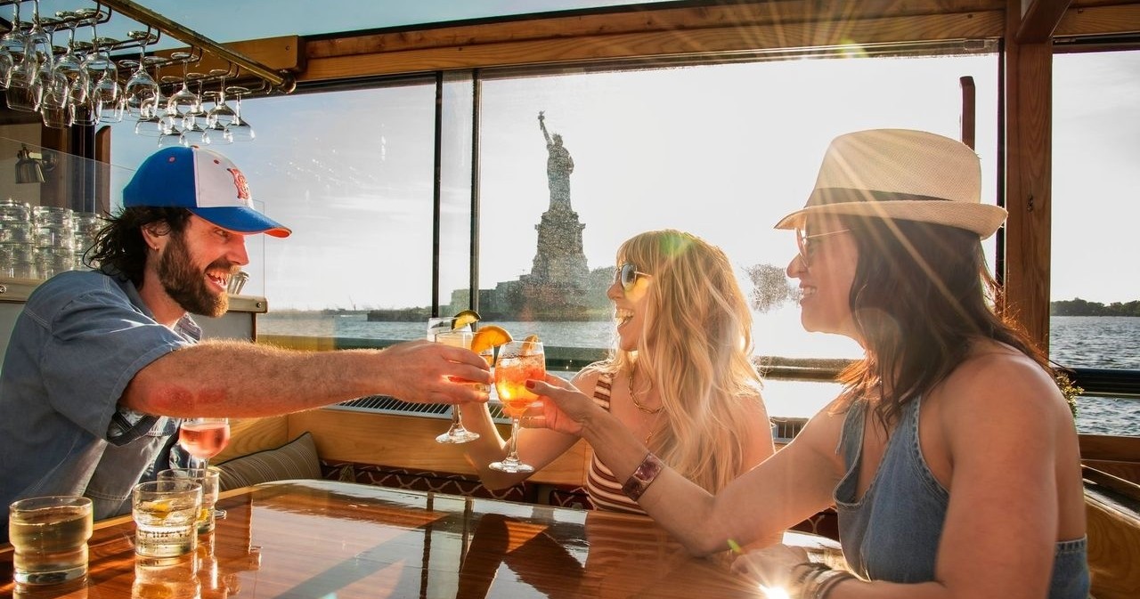 Three people toasting drinks on a boat near the Statue of Liberty at sunset.