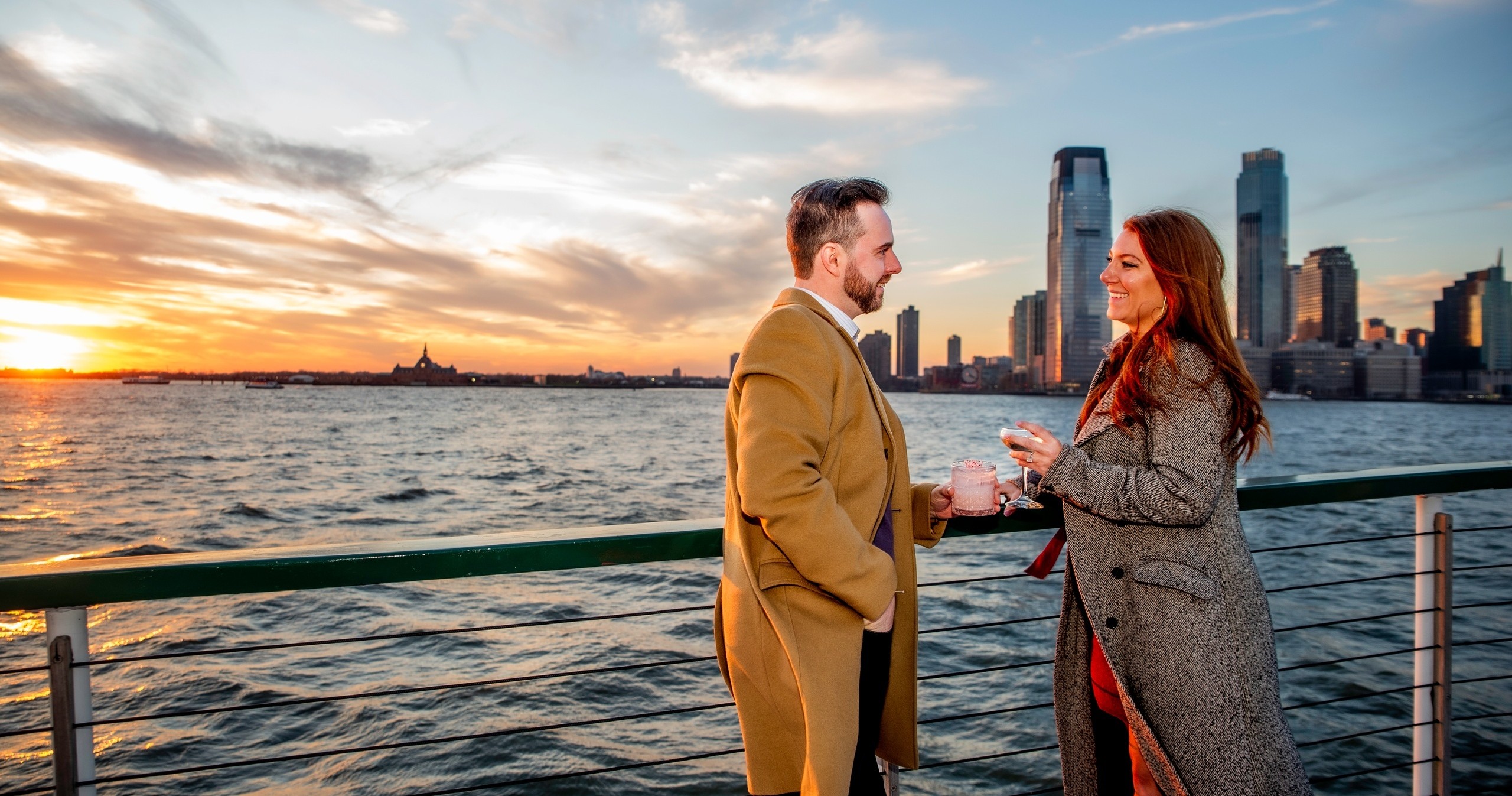 Two people smiling, holding cups by waterfront at sunset, city skyline in background.