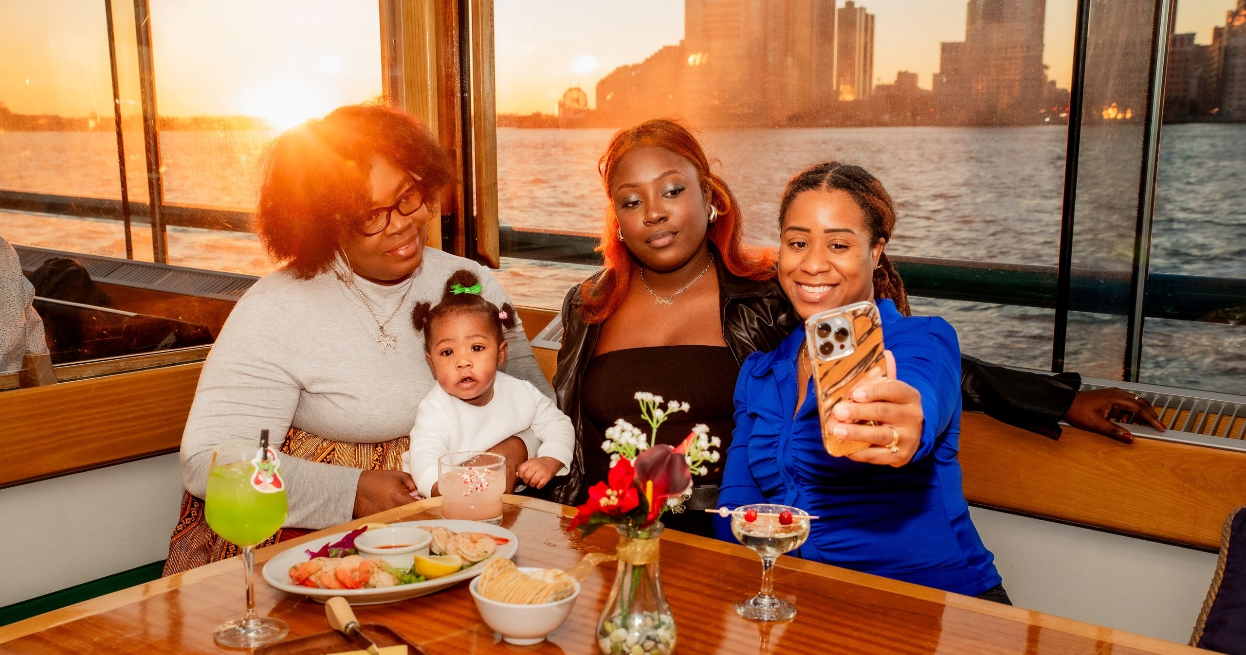 Three women and a baby pose for a selfie on a boat with a city skyline at sunset.