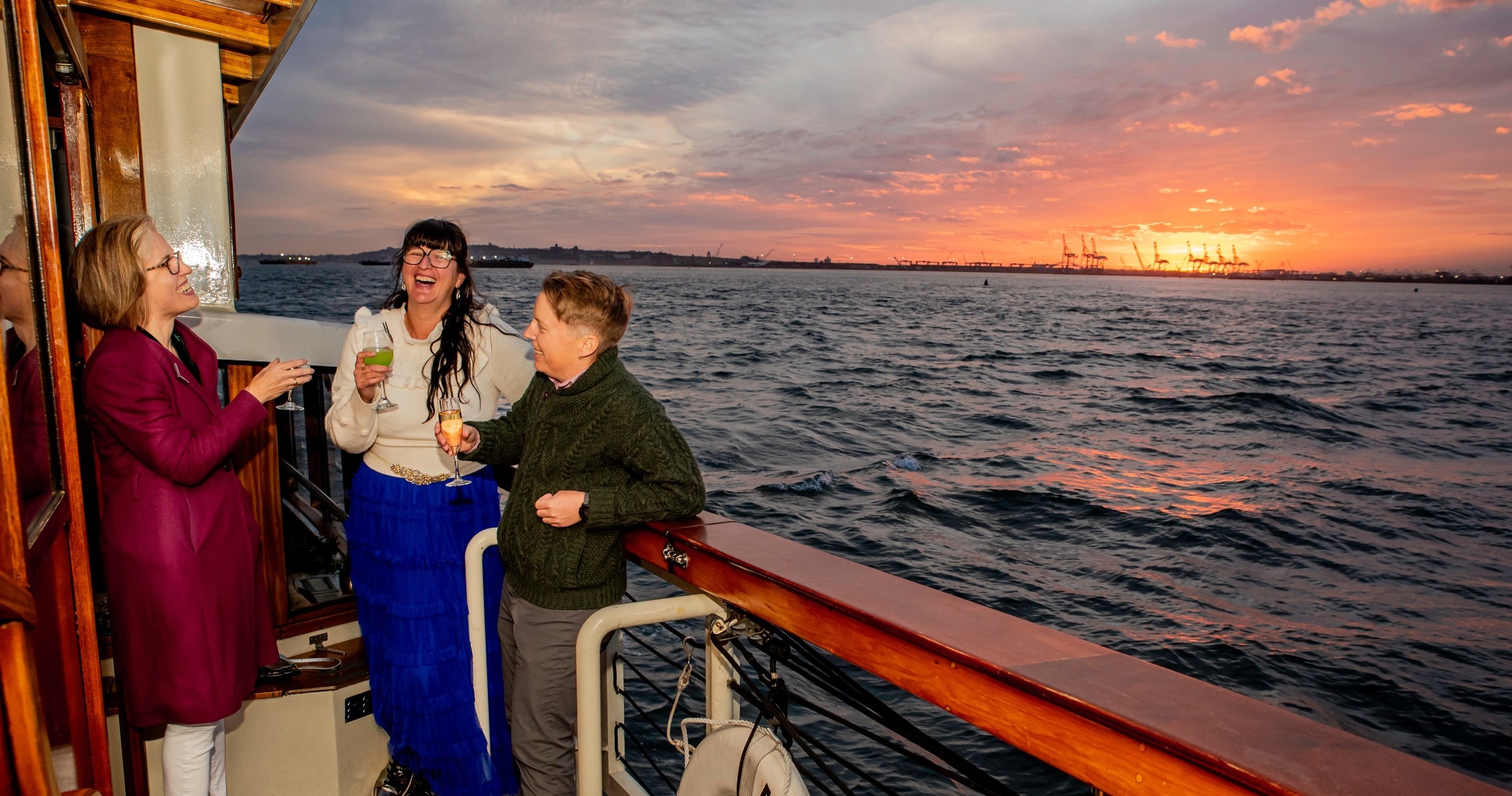 Three people laughing on a boat, with a sunset over the ocean in the background.
