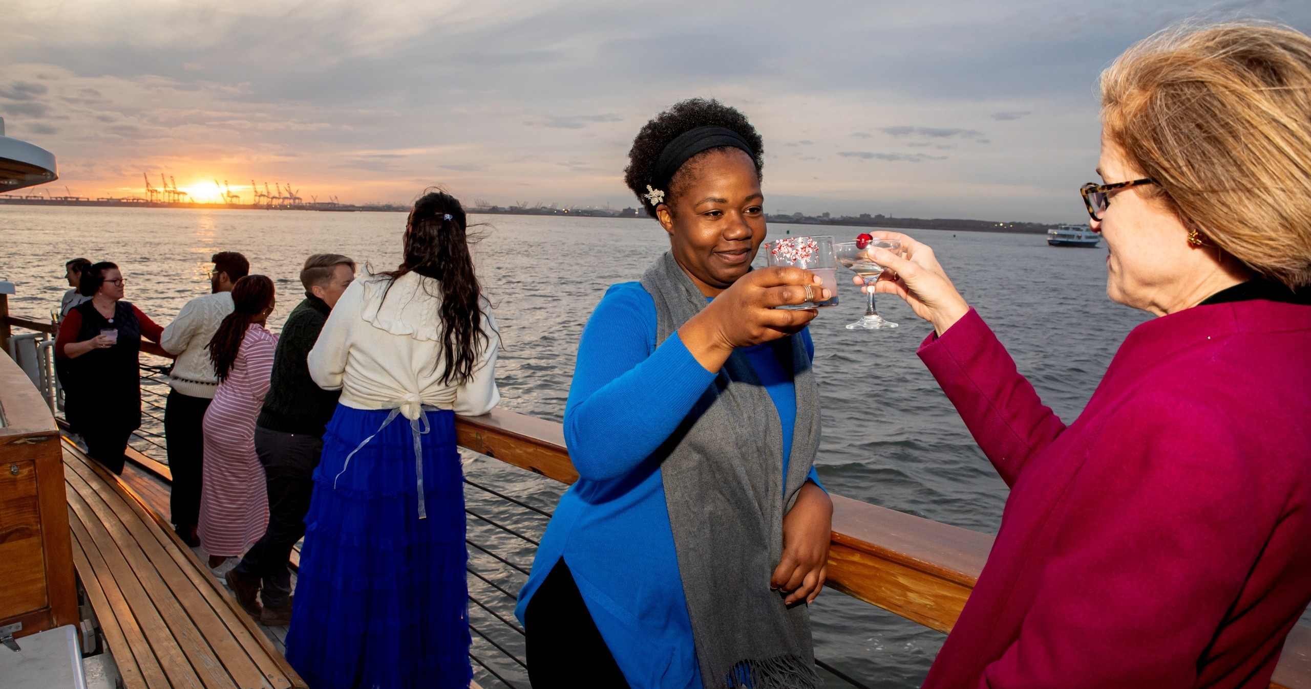 People on a boat deck toasting as the sun sets over the water.