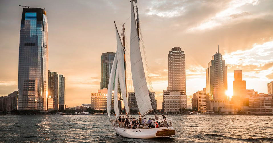Sailboat on water with skyscrapers in background at sunset.