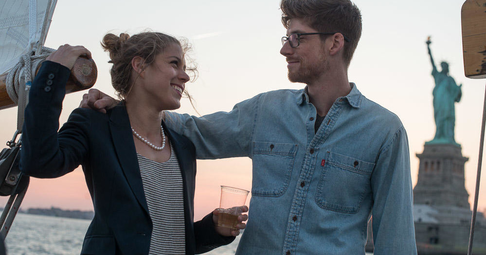Two people smiling on a boat by the Statue of Liberty at sunset.