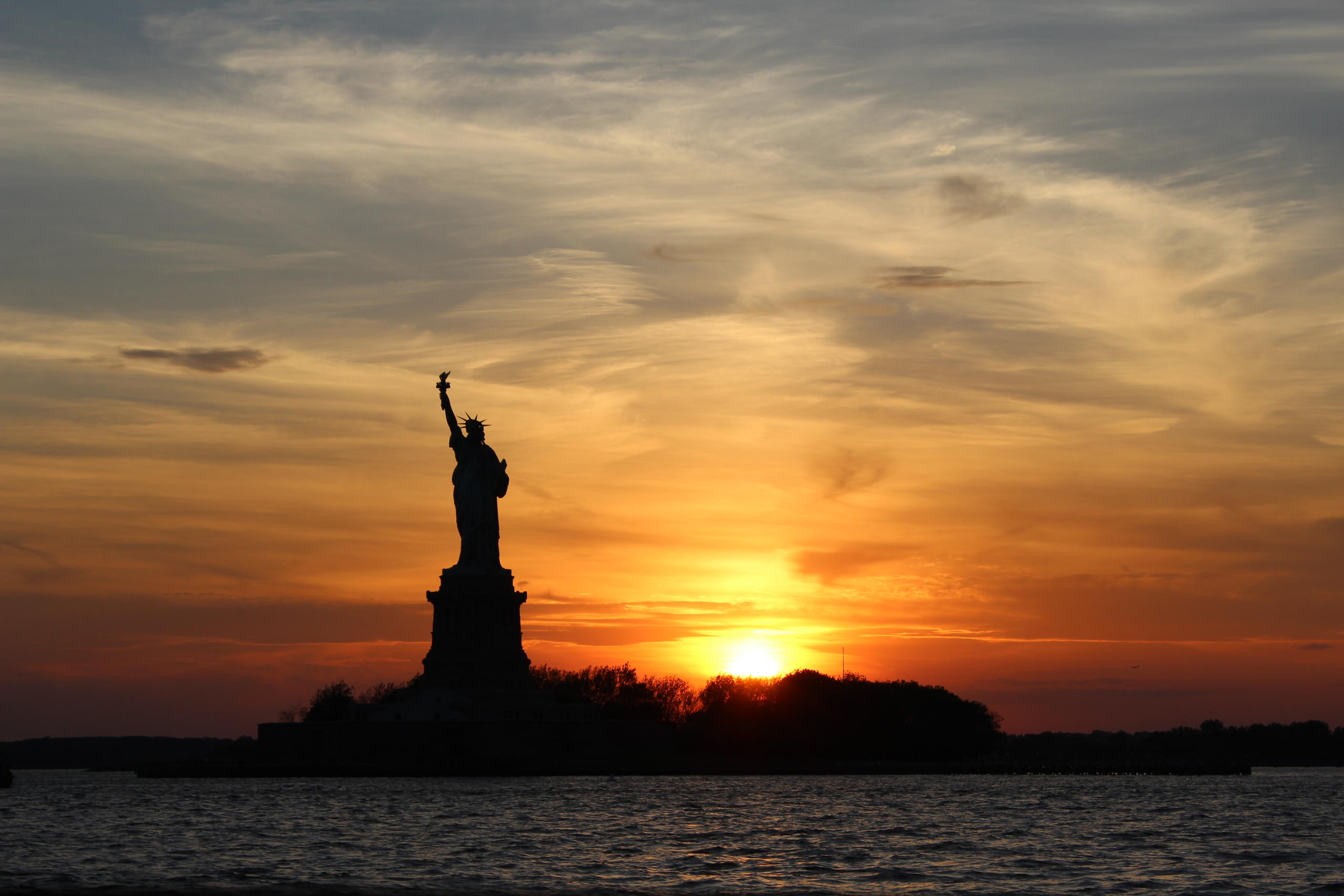Silhouette of Statue of Liberty against a sunset with scattered clouds.