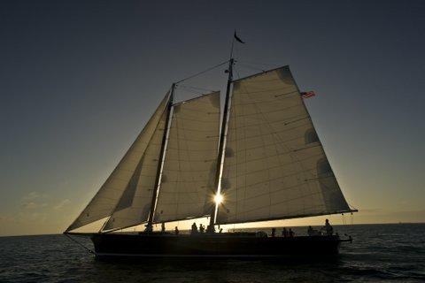 Silhouetted sailboat with sun peeking through sails on the water at sunset.
