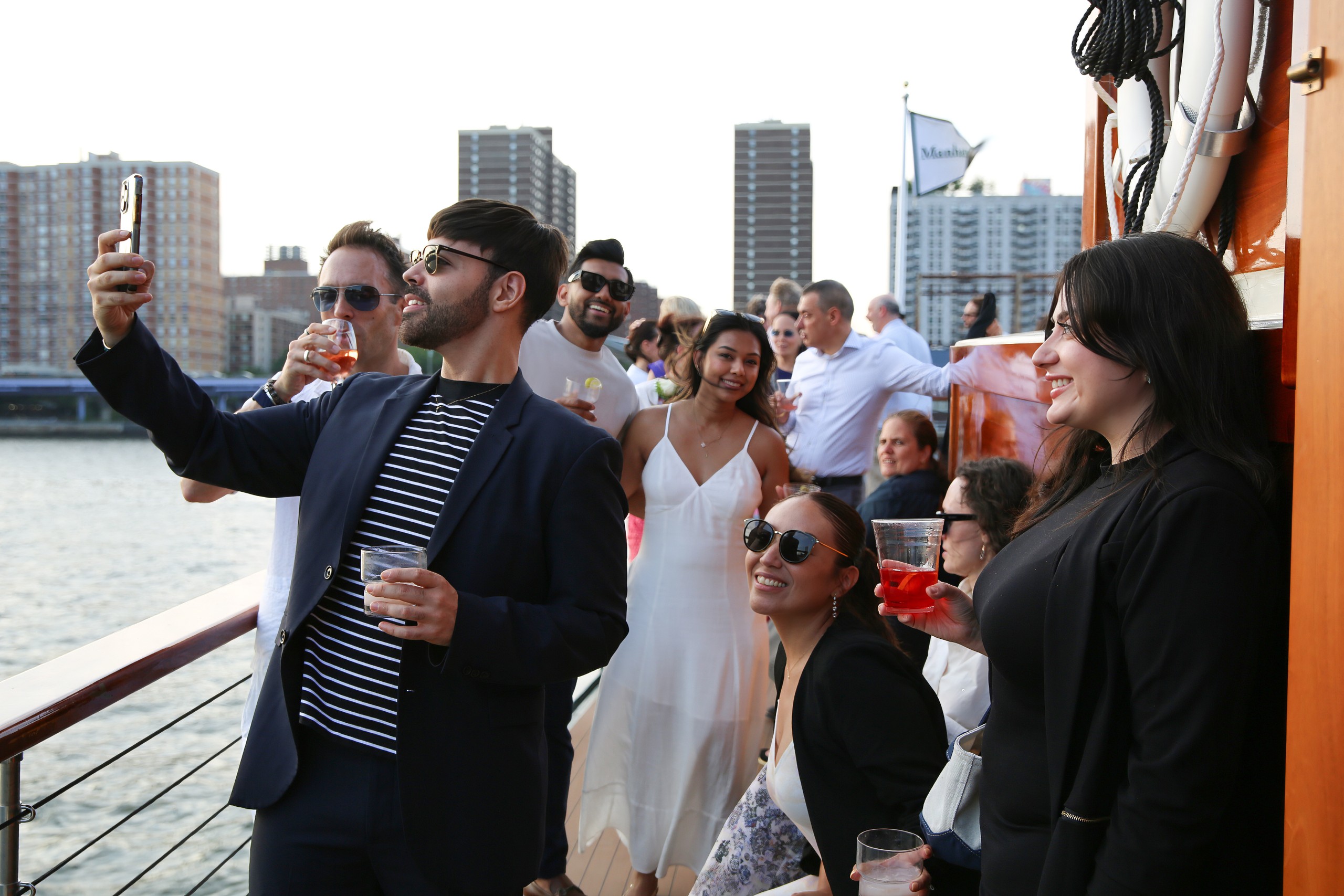 Group of people on a boat taking selfies, holding drinks, city buildings in background.