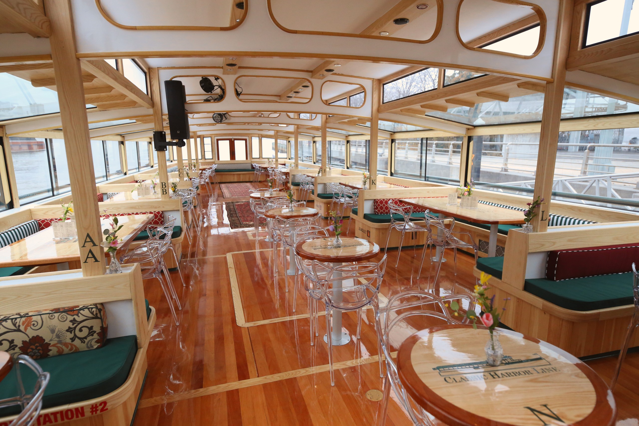 Interior of a wooden riverboat with tables, clear chairs, and large windows.