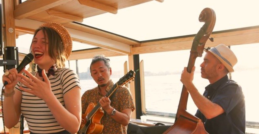 Woman singing with two musicians on a boat, one playing guitar and the other playing double bass.