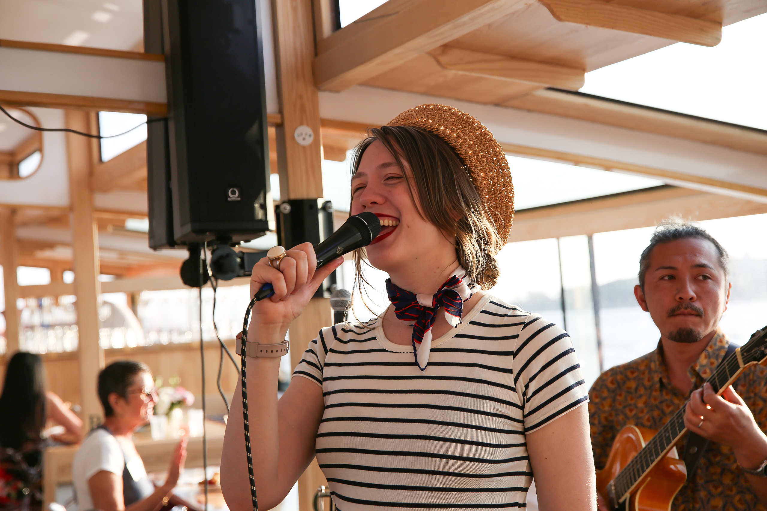 Woman singing into a microphone, man playing guitar, sunny indoor setting.