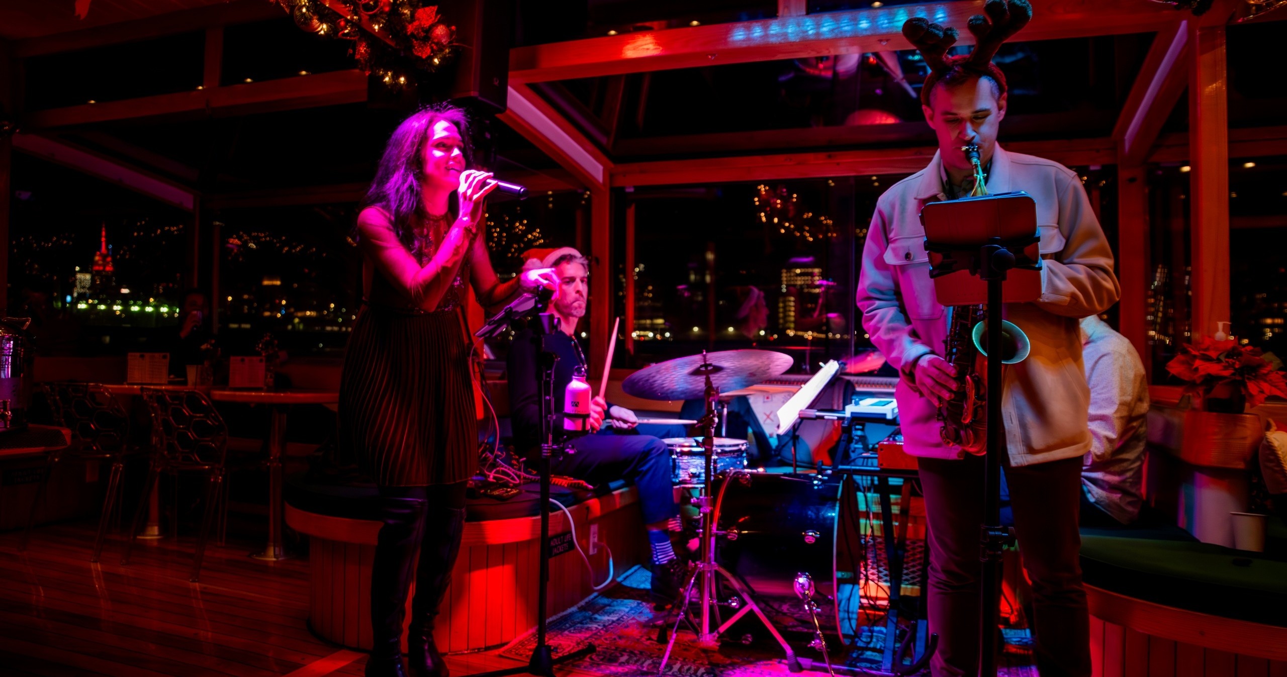 A band performs on stage with singer, drummer, and saxophonist with reindeer antlers, in a dimly lit venue.