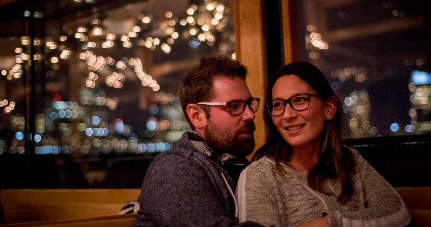 Couple seated inside with city lights reflecting in the window at night.
