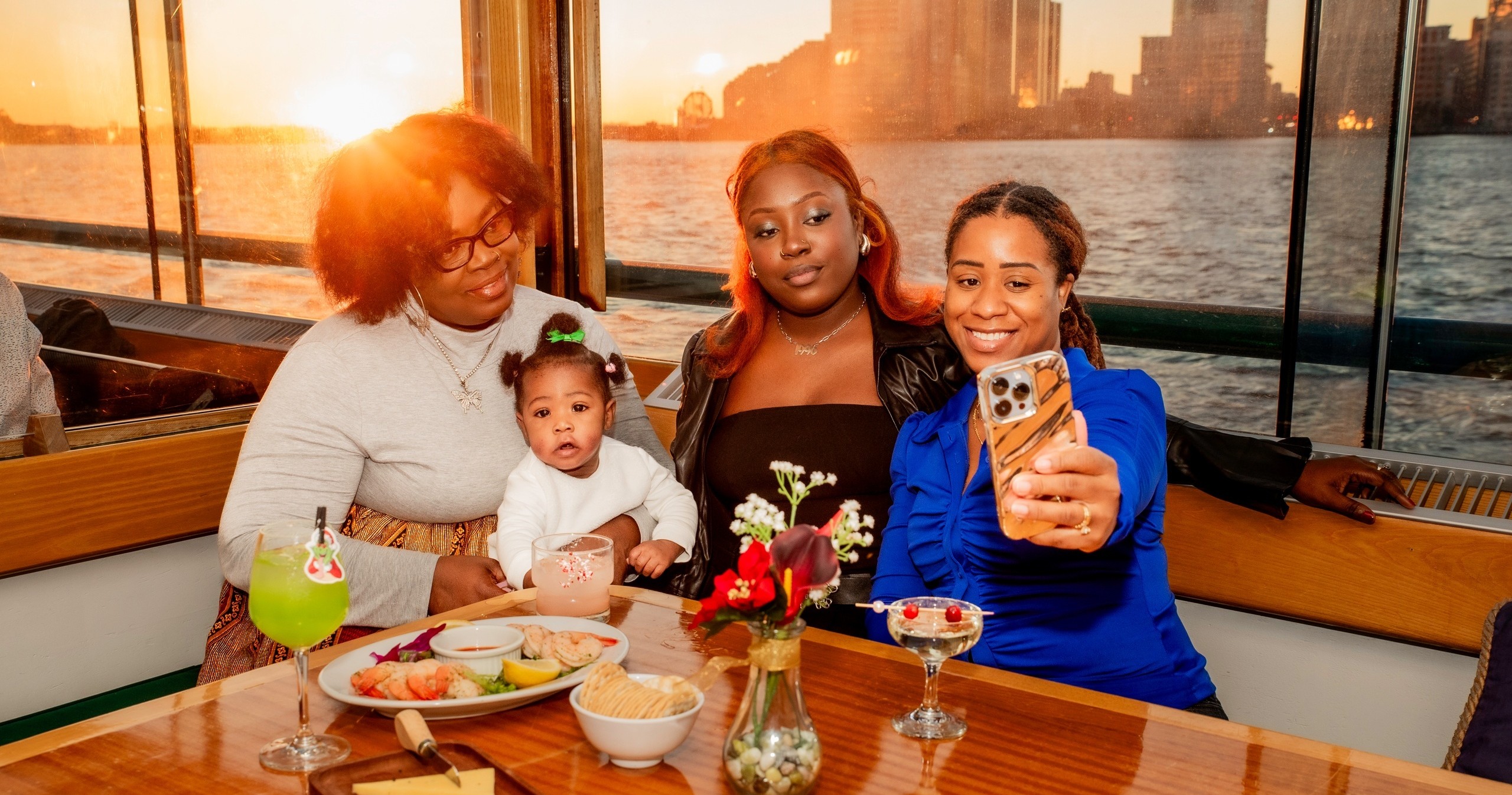 Three women and a baby pose for a selfie at sunset on a boat.