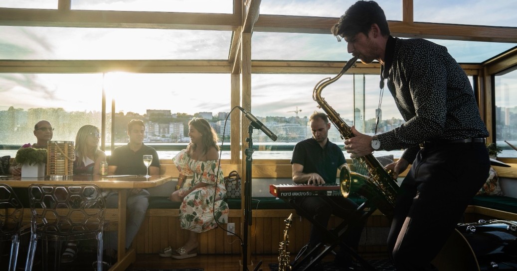 Saxophonist plays to seated audience in a sunlit room with a cityscape view.