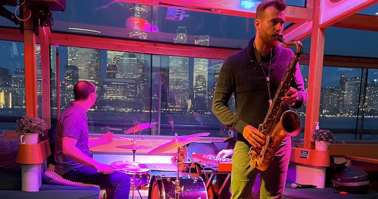 Musician playing saxophone with drummer, city skyline in background.