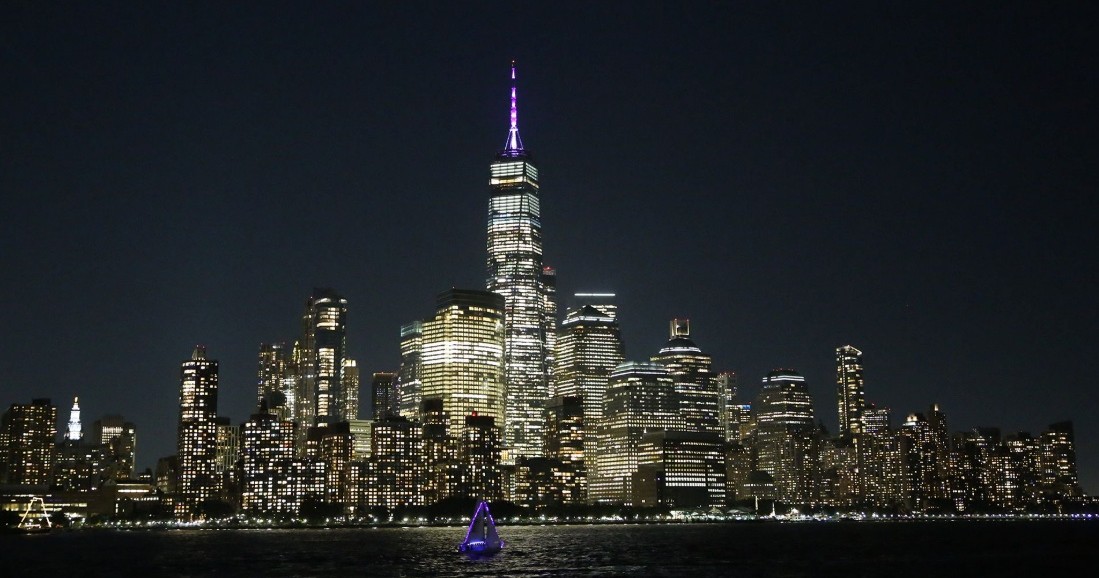 New York City skyline at night with lit purple sailboat on water.