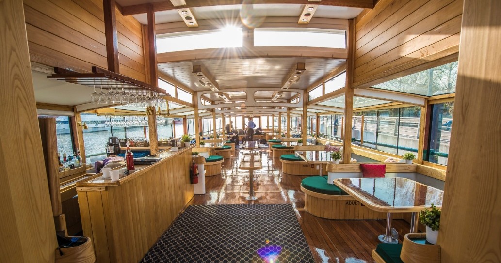 Wood-paneled boat interior with tables and windows overlooking water.
