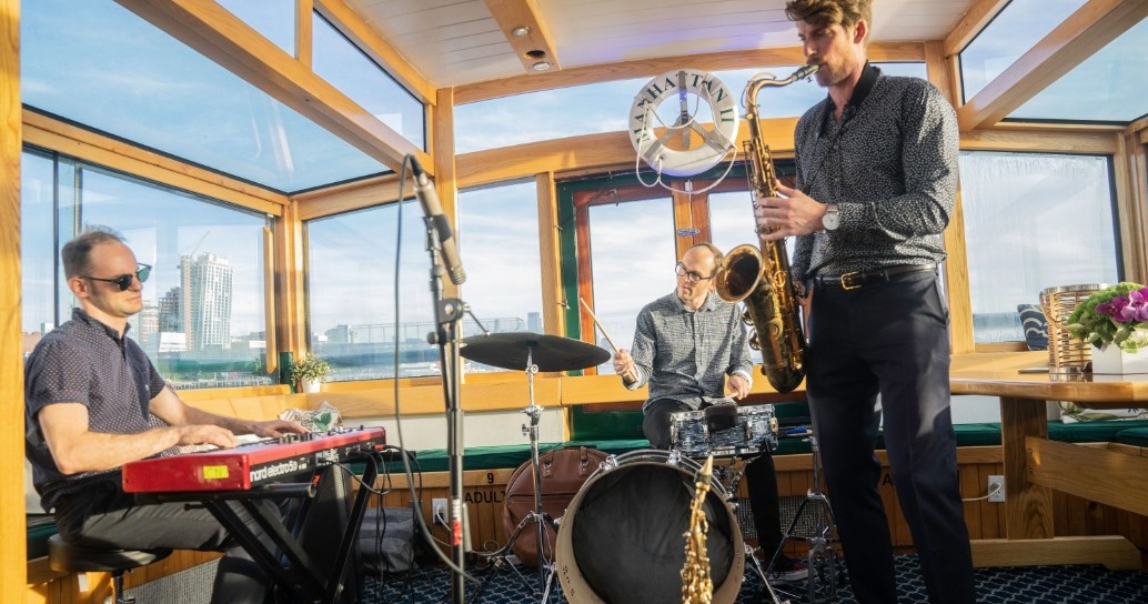 Three musicians performing on a boat: one on keyboard, one on drums, and one on saxophone.