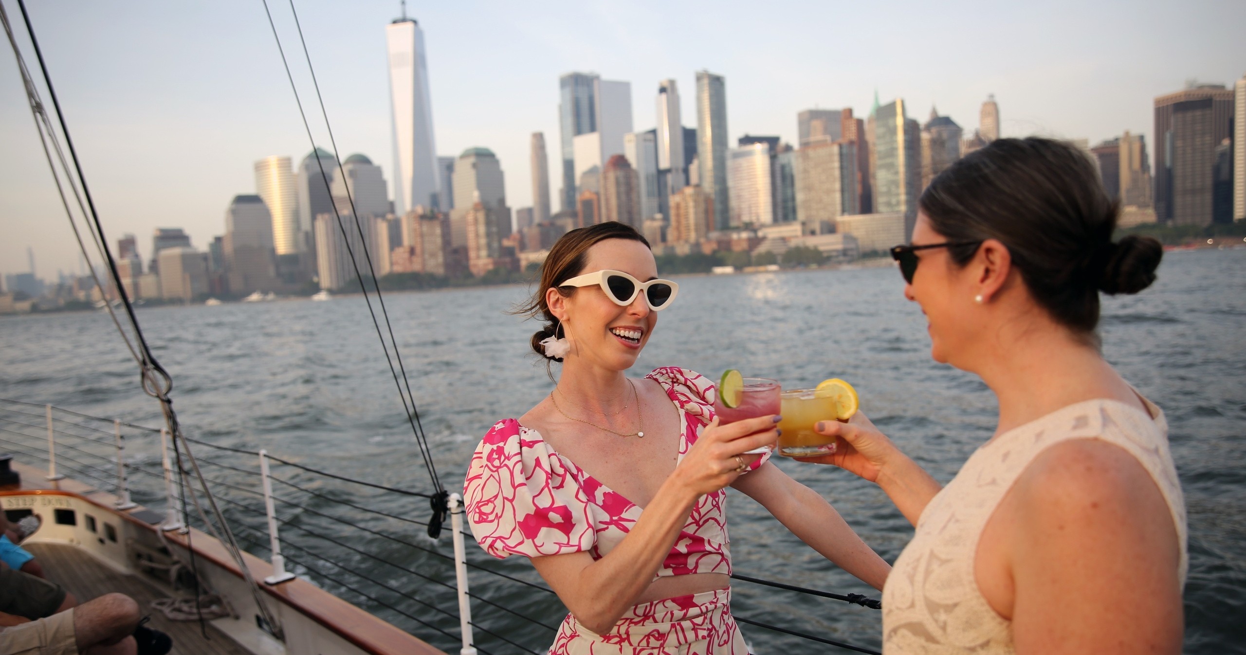 Two women toasting drinks on a boat with city skyline in background.