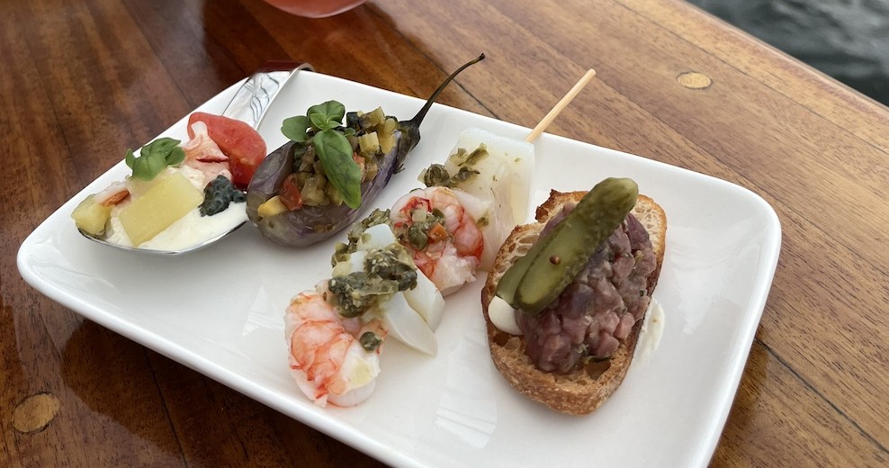 Gourmet appetizers on a white plate, including shrimp, eggplant, and a crostini with pickle on wooden table.