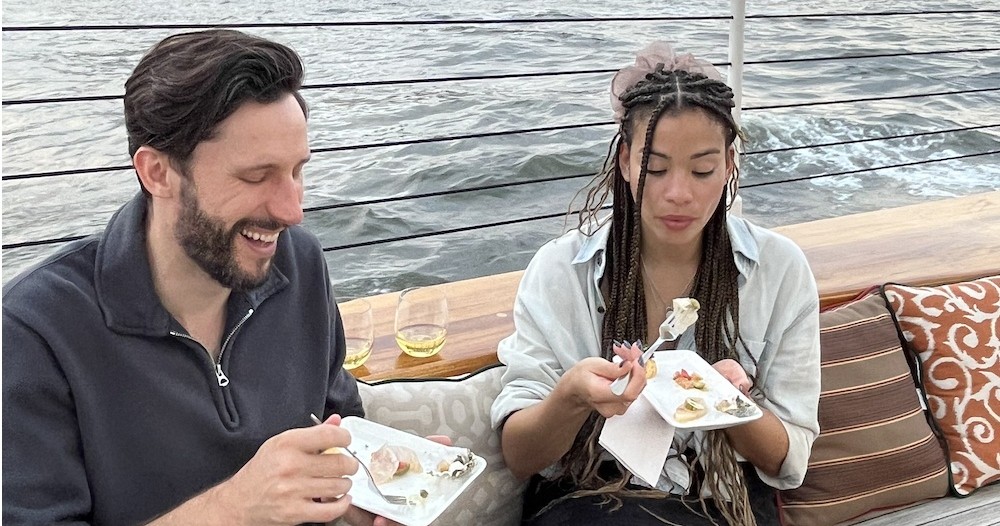 Two people eating on a boat, holding plates with food, water in the background.
