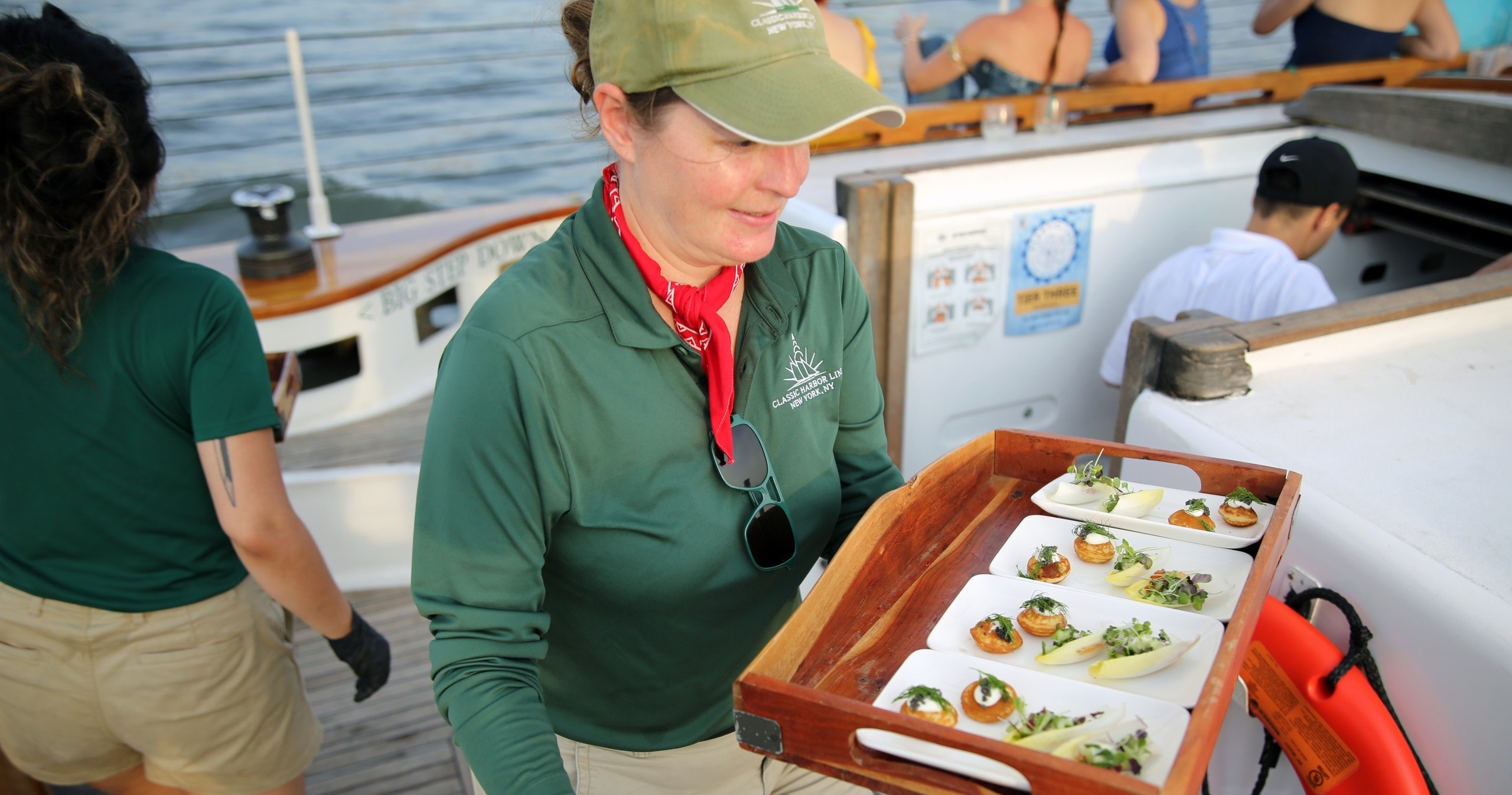 Server in green uniform carries tray of appetizers on a boat deck.