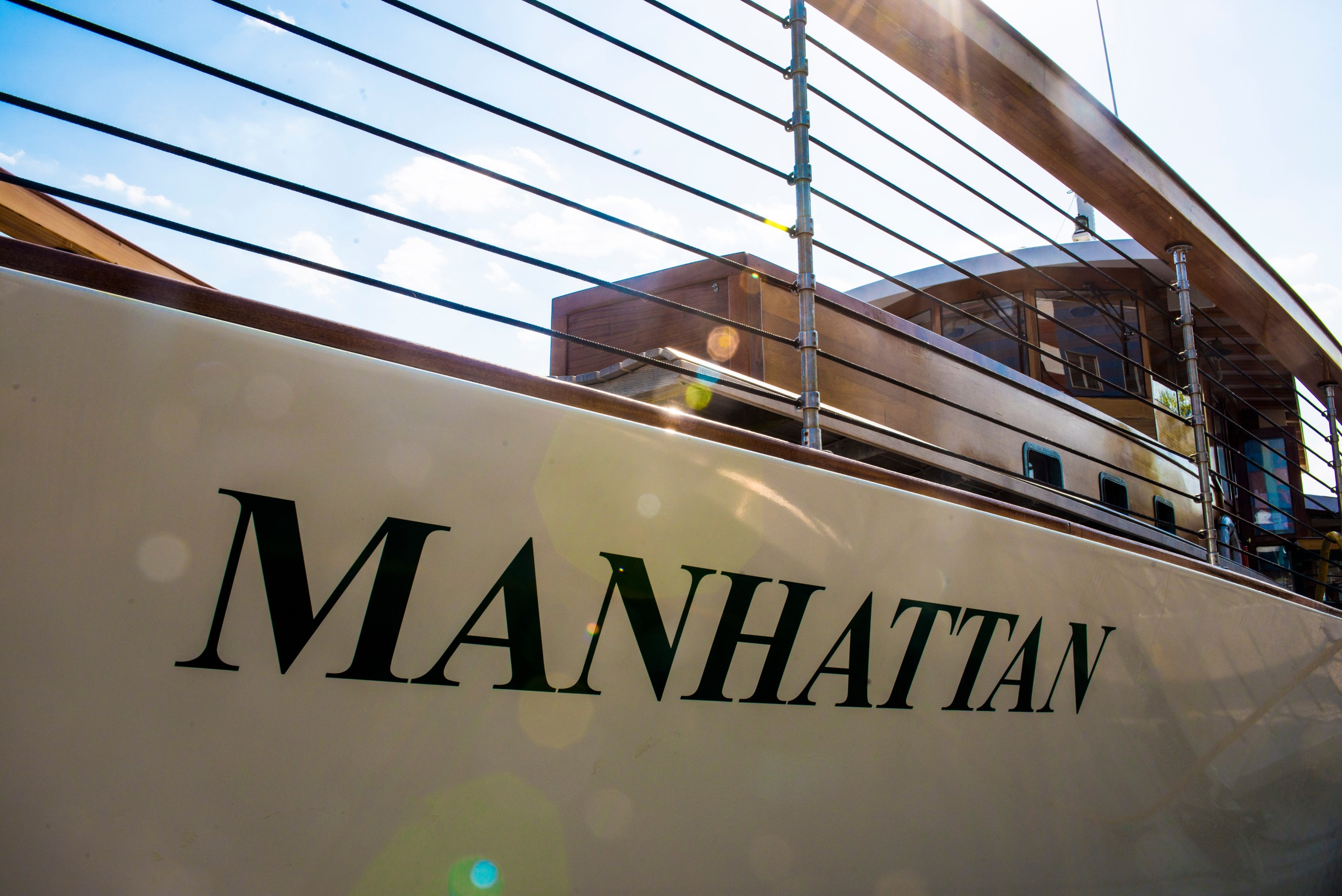 Close-up of a yacht side with 'MANHATTAN' written on it, railing above, and sun glare in the image.