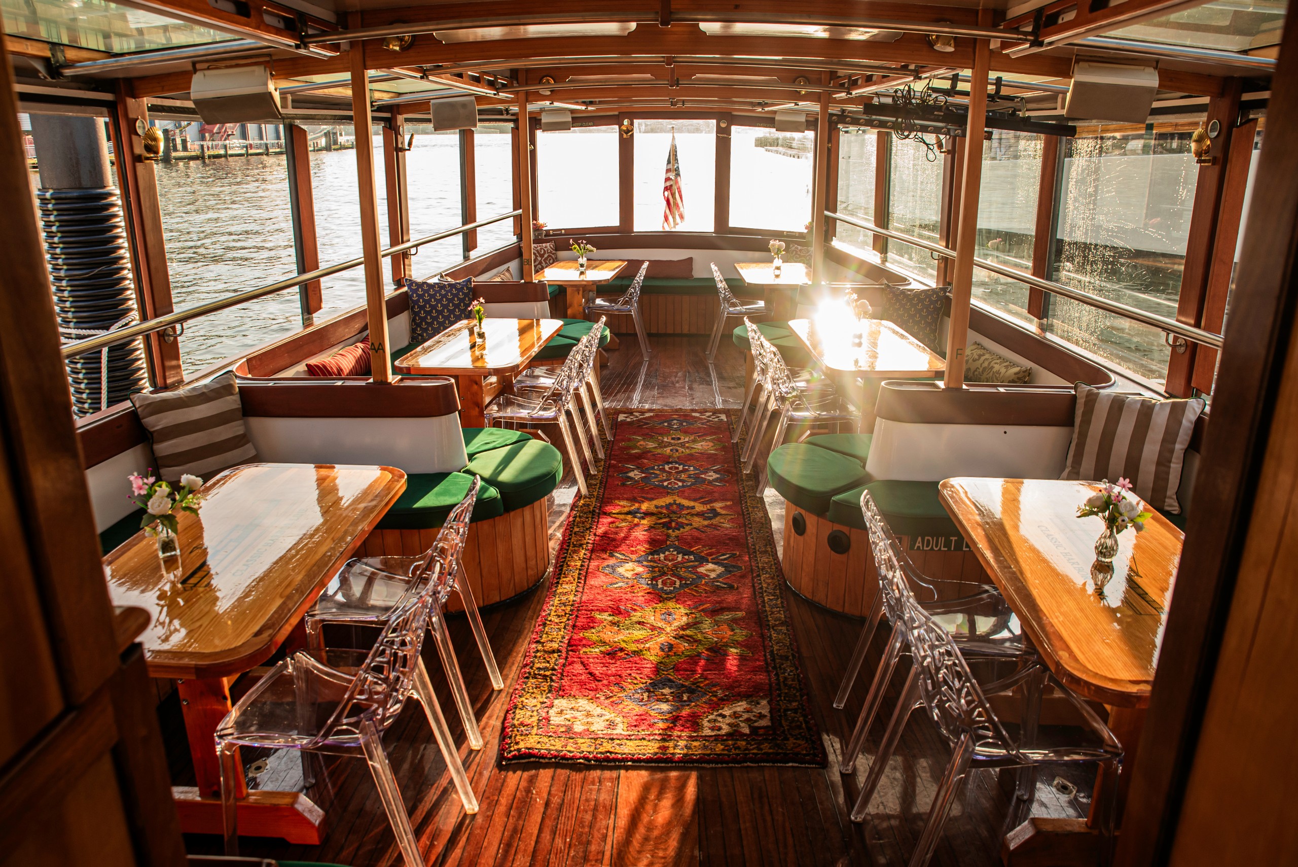 Sunlit boat interior with tables, chairs, and floral arrangements on a red rug, view of water and American flag.