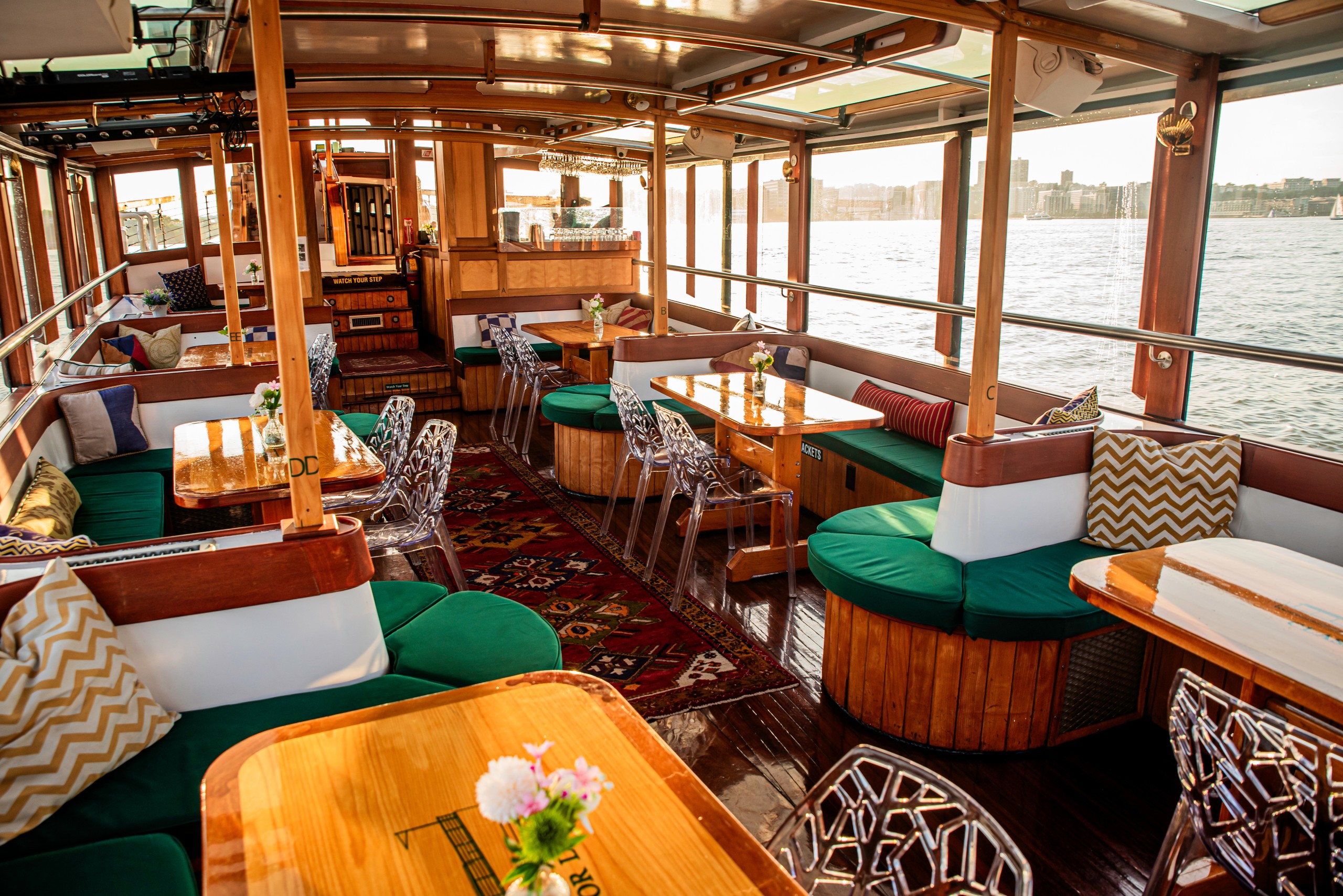 Cozy boat lounge with wooden tables, green seating, and large windows overlooking the water.