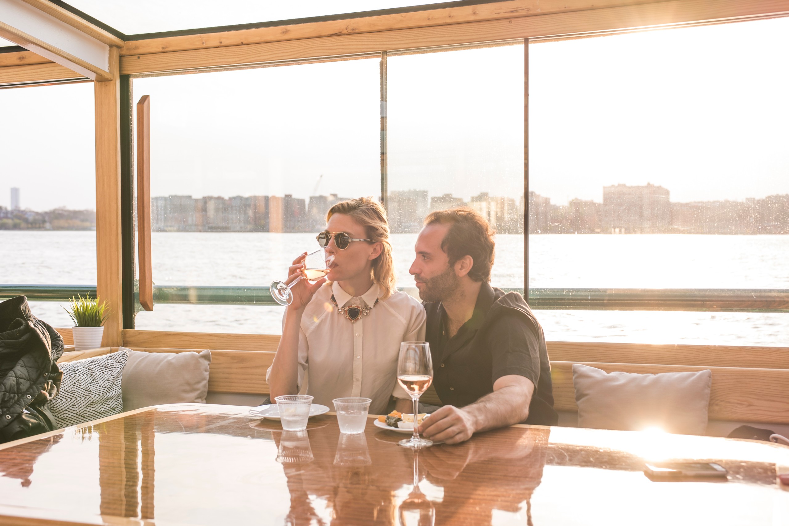 Two people on a boat, enjoying drinks at sunset with city skyline in the background.