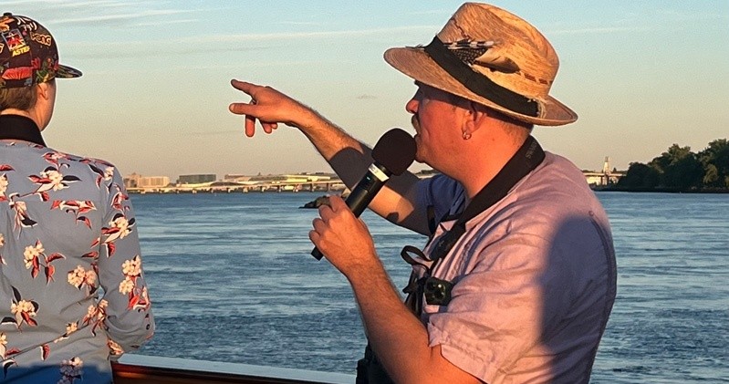 Man with microphone and hat pointing by the water.