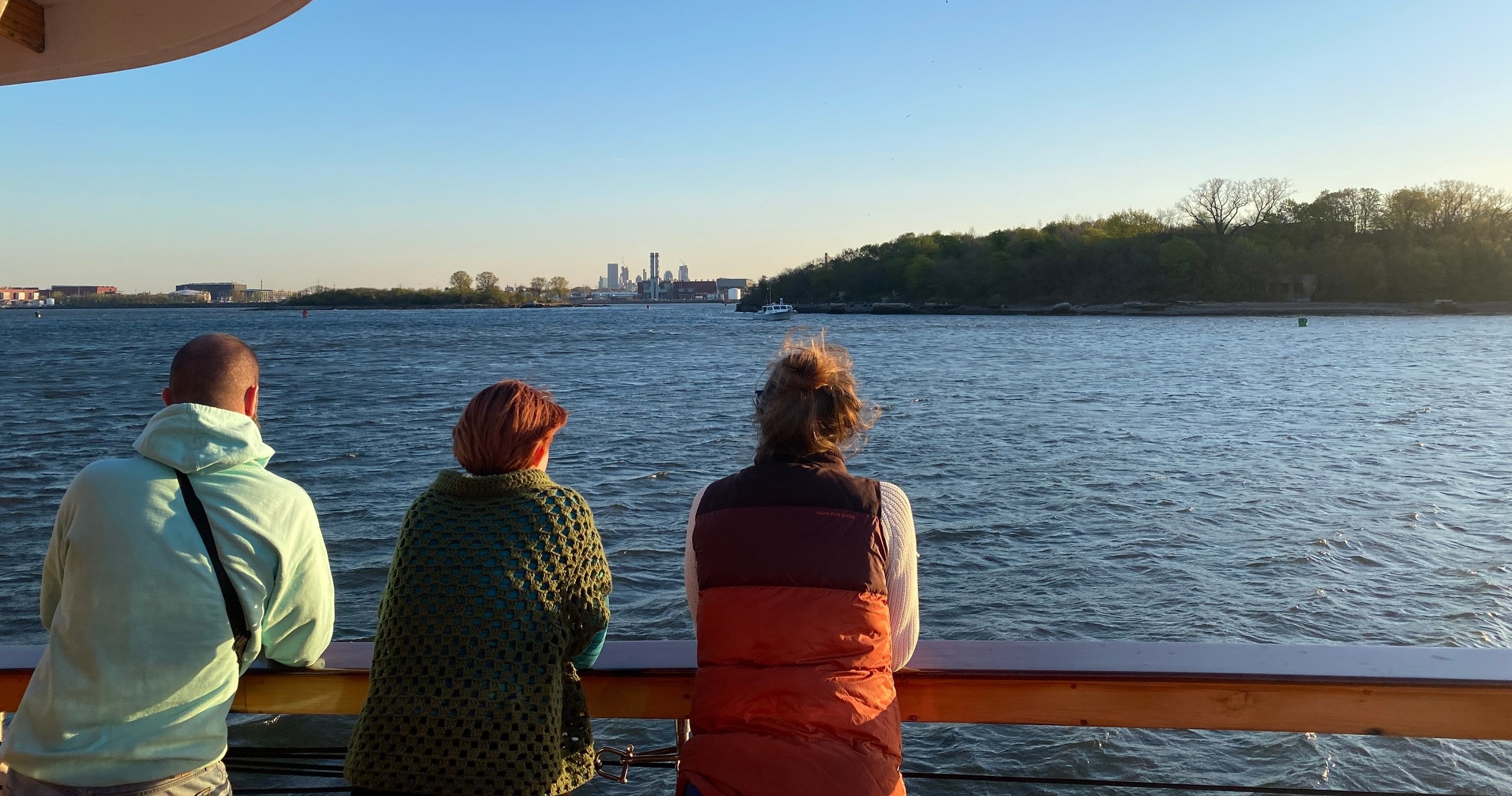 Three people look at water from a ferry, city skyline in the background during sunset.