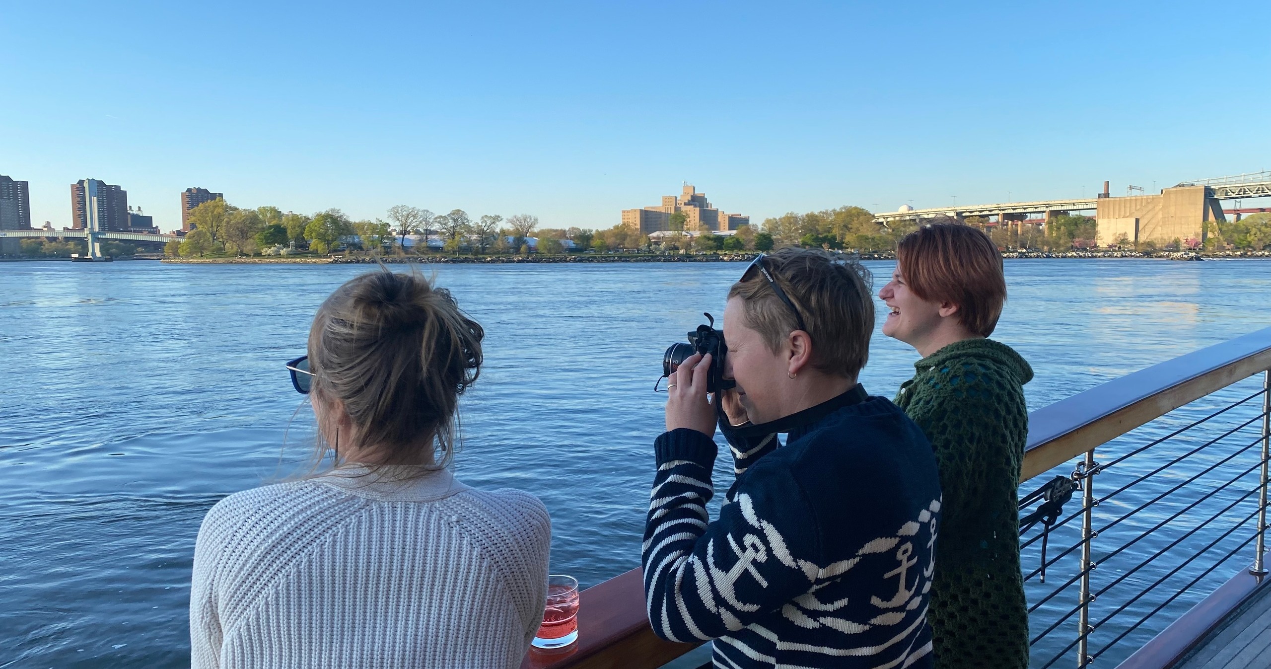 Three people on a boat, one taking photos, with a river and city skyline in the background.