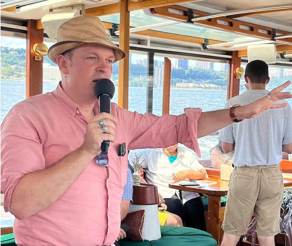 Man in pink shirt and hat speaks into a microphone on a boat.