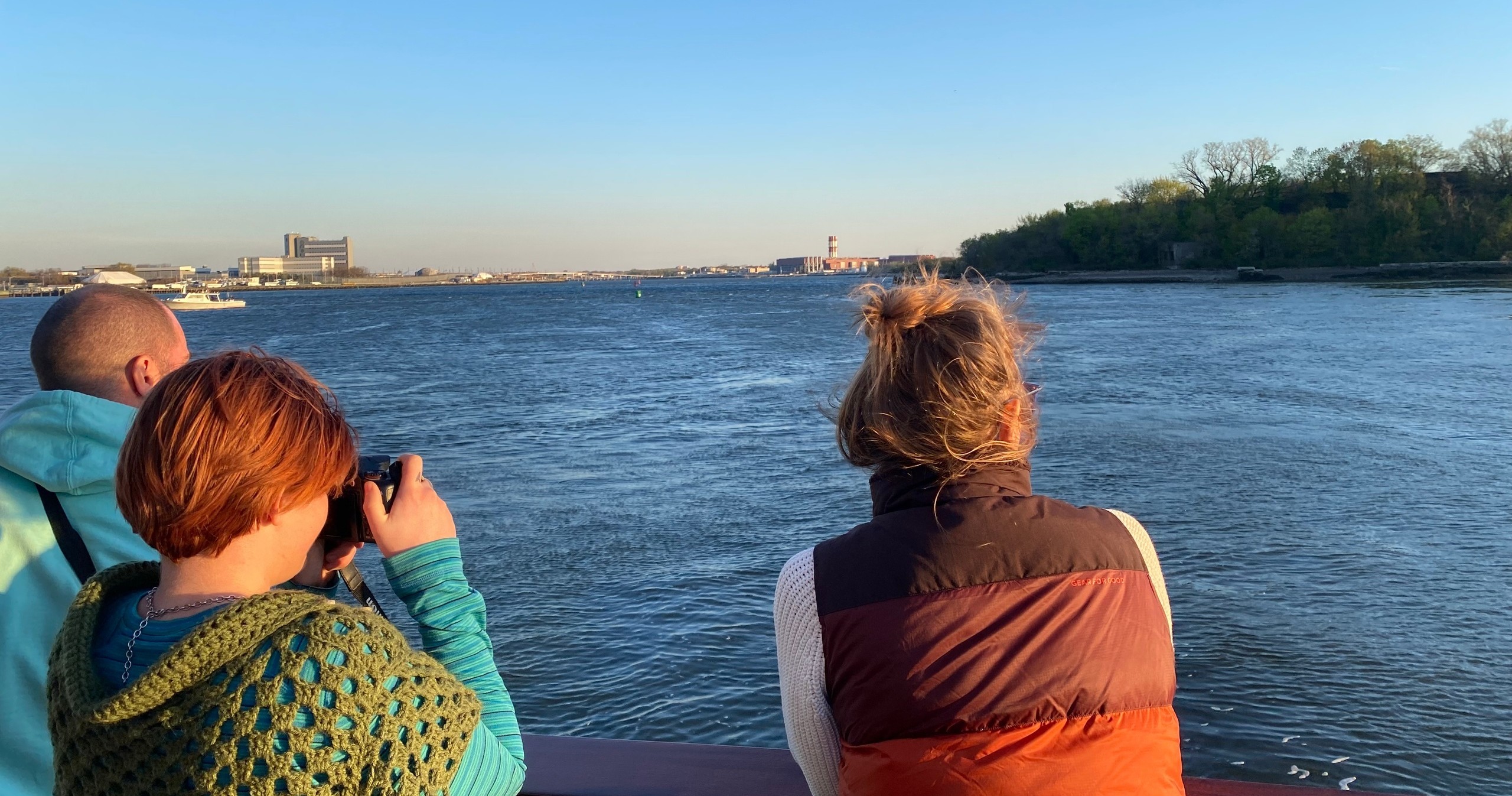 Three people on a boat observing a river view under clear skies.