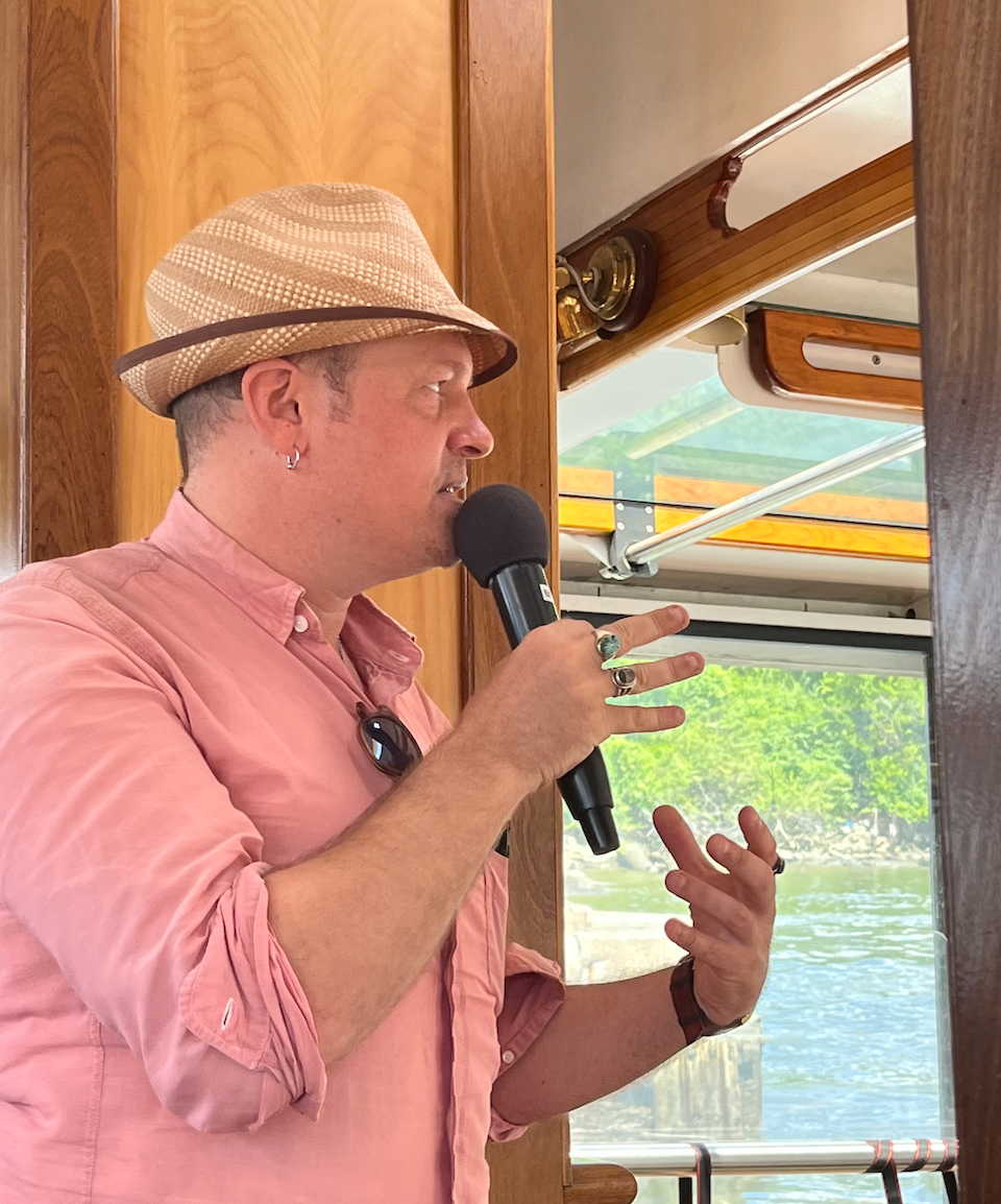 Person speaking into a microphone on a boat, wearing a hat and pink shirt.