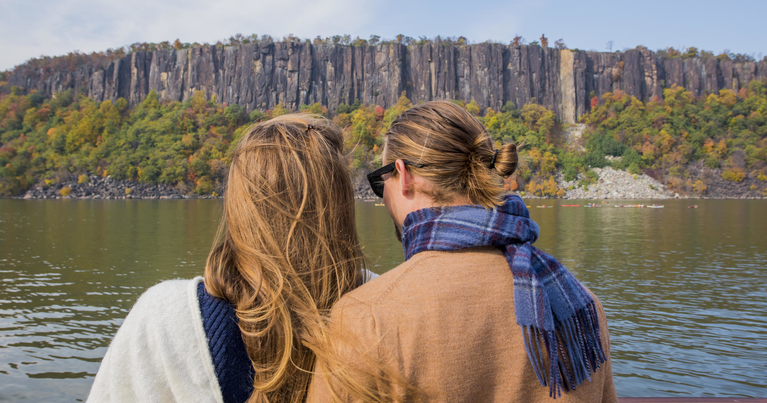 Two people by a river view tall cliffs and autumn trees.