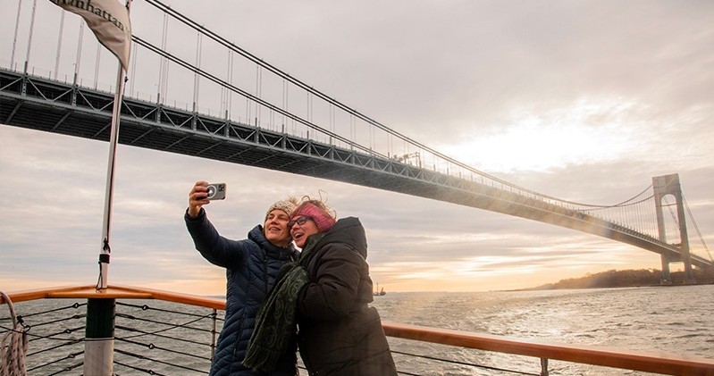 Two people taking a selfie on a boat near a large suspension bridge.